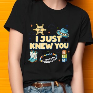May include: A black t-shirt featuring a graphic print with the text I JUST KNEW YOU, a yellow sheriff badge with TO INFINITY AND BEYOND, a blue cowboy hat, light blue boots, a beaded bracelet with I'VE GOT A FRIEND IN ME, and three toy blocks.