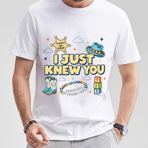 May include: A white short-sleeved t-shirt featuring a graphic print with the text I JUST KNEW YOU, a yellow sheriff star badge with TO INFINITY AND BEYOND, a blue cowboy hat, light blue boots, a beaded bracelet with YOU'VE GOT A FRIEND IN ME, and blocks.