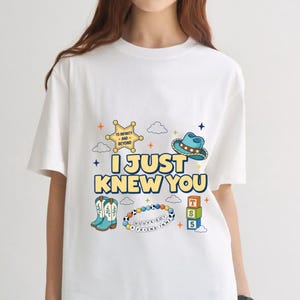 May include: A white short-sleeve t-shirt featuring a graphic print with the text I JUST KNEW YOU, a yellow sheriff badge, a blue cowboy hat, light blue cowboy boots, a colorful beaded bracelet, and three stacked blocks labeled T, S, and 5.