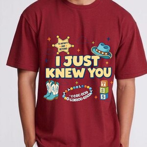May include: A maroon short-sleeved t-shirt featuring the yellow text I JUST KNEW YOU. Graphics include a yellow sheriff badge with TO INFINITY AND BEYOND, a blue cowboy hat, light blue boots, a colorful beaded bracelet, and three stacked alphabet blocks.