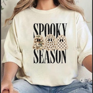 May include: A cream-colored t-shirt featuring the black text SPOOKY SEASON. Below the word SPOOKY are three ghost illustrations with different patterns: one with camouflage, one with a leopard print, and one with a tan and white checkerboard pattern.