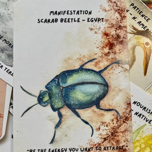 Könnte beinhalten: Eine rechteckige Karte mit einer Aquarellillustration eines blau-grünen Skarabäus-Käfers. Der Text lautet: MANIFESTATION SCARAB BEETLE - EGYPT und BE THE ENERGY YOU WANT TO ATTRACT. - UNKNOWN. Der Hintergrund enthält braune, texturierte Farbspritzer.