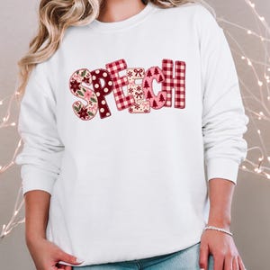 May include: A white long-sleeve sweatshirt featuring the word SPEECH in large, block-style letters. Each letter has a unique holiday-themed pattern, including red and white buffalo plaid, floral prints, snowflakes, bows, and Christmas trees.