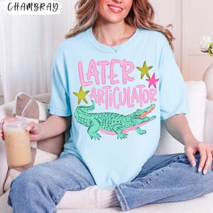 May include: A light blue t-shirt featuring a graphic of a green alligator and the pink text "LATER ARTICULATOR" with yellow and pink stars. The shirt has a crew neckline and short sleeves, showcasing a casual design with a playful animal illustration.