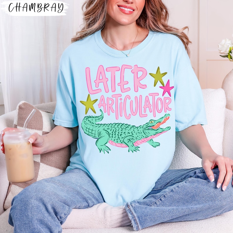 May include: A light blue t-shirt featuring a graphic of a green alligator and the pink text "LATER ARTICULATOR" with yellow and pink stars. The shirt has a crew neckline and short sleeves, showcasing a casual design with a playful animal illustration.