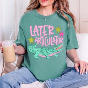 May include: A sage green short-sleeve t-shirt featuring the pink text "LATER ARTICULATOR" above a green alligator illustration. The design includes two yellow stars and one pink star. The shirt has a crew neckline and a relaxed fit.