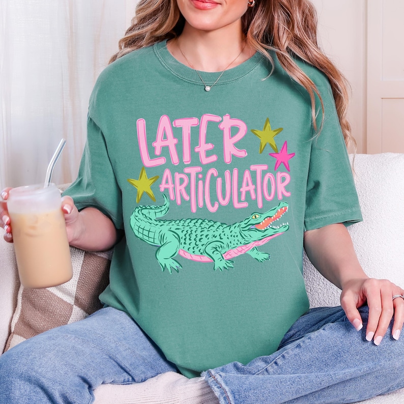 May include: A sage green short-sleeve t-shirt featuring the pink text "LATER ARTICULATOR" above a green alligator illustration. The design includes two yellow stars and one pink star. The shirt has a crew neckline and a relaxed fit.