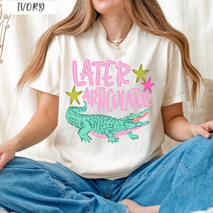 May include: An ivory short-sleeve t-shirt featuring a graphic print of a green alligator with a pink underbelly. Above the alligator, the text reads "LATER ARTICULATOR" in pink letters, accompanied by one yellow star and one pink star graphic.
