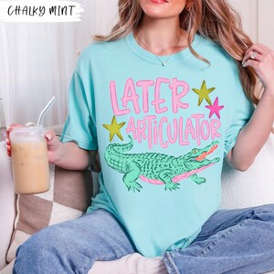 May include: A chalky mint t-shirt featuring a graphic of a green alligator and the pink text "LATER ARTICULATOR" with yellow and pink stars. The design is centered on the front of the shirt, which is worn with blue denim jeans.