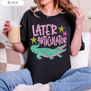 May include: A black t-shirt featuring a graphic print of a green alligator and the text "LATER ARTICULATOR" in pink lettering. The design includes two small stars in yellow and pink, positioned above the alligator on the front of the shirt.