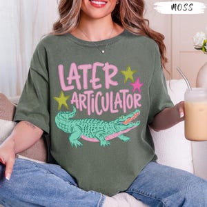 May include: A moss green short-sleeve t-shirt featuring the pink text "LATER ARTICULATOR" above a green alligator illustration. The design includes two yellow stars and one pink star. The shirt has a crew neckline and a relaxed, loose fit.