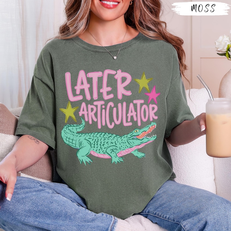 May include: A moss green short-sleeve t-shirt featuring the pink text "LATER ARTICULATOR" above a green alligator illustration. The design includes two yellow stars and one pink star. The shirt has a crew neckline and a relaxed, loose fit.