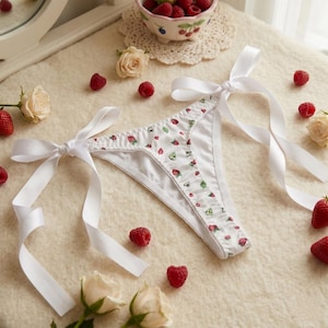 May include: White thong underwear with a strawberry and leaf pattern, tied with white satin ribbons. Fresh raspberries and roses are scattered around the lingerie, creating a delicate and feminine aesthetic.