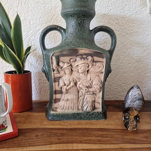 Op de afbeelding: Een vintage groene keramische vaas met twee handvatten en een gedetailleerd reliëf van figuren in historische kleding. De vaas heeft een getextureerd oppervlak en een brede, cilindrische hals. Het reliëf is lichter van kleur.