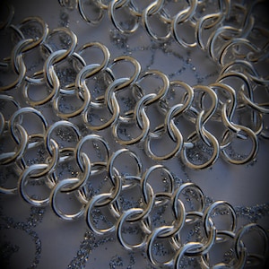 May include: Close-up of shiny silver chainmail links arranged in rows. The links are interlocked, creating a flexible, textured surface. The background is a light, glittery surface, enhancing the metallic sheen of the chainmail.