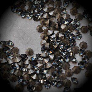May include: A close-up of a large quantity of dark grey and black rhinestones. The rhinestones are scattered on a white surface.