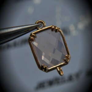 Puede incluir: Un colgante dorado con una piedra preciosa rosa claro facetada y transparente de forma cuadrada. El colgante tiene un bucle en la parte superior para sujetarlo a una cadena.