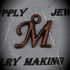 Puede incluir: Un abalorio de metal de color cobre en forma de letra M con un bucle en la parte superior. La letra está en una fuente cursiva y está sobre un fondo blanco con texto negro que dice "Supply Jewelry Making".