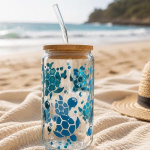 May include: A clear glass tumbler with a bamboo lid and a glass straw. The tumbler is decorated with blue sea turtle designs and glitter. The tumbler is sitting on a white blanket on a sandy beach, with a straw hat in the background.