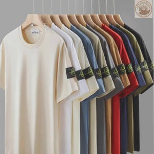 May include: A collection of short-sleeved t-shirts in various colors, including cream, white, blue, and red, hanging on wooden hangers. Each shirt features a small, square patch with a circular logo on the left sleeve.