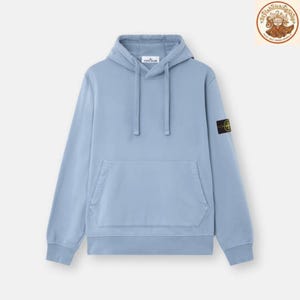 Peut inclure: Un sweat à capuche bleu clair avec une poche avant et une capuche à cordon. Un petit écusson noir et jaune est visible sur la manche gauche. Le nom de la marque est visible sur la capuche.