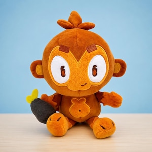 May include: A plush toy monkey with large white-rimmed eyes, brown fur, and a small, smiling mouth. The monkey is holding a black and yellow object. The toy is sitting on a light blue background.