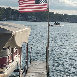 Puede incluir: Una bandera de los Estados Unidos ondea en un poste de metal al final de un muelle de madera que se extiende hacia un lago. A la izquierda, se ve parte de un barco pontón con un toldo beige. A lo lejos, un pequeño barco se mueve por el agua.