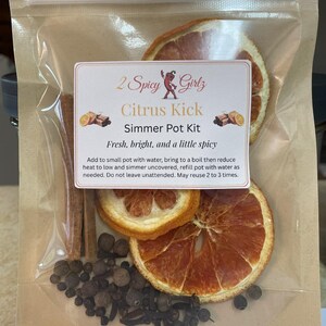 Könnte beinhalten: Ein klarer Beutel mit einem Zitrus-Simmer-Pot-Kit. Das Kit enthält getrocknete Orangenscheiben, Zimtstangen, ganze Pimentkörner und Nelken. Auf dem Etikett steht "2 Spicy Girlz Citrus Kick Simmer Pot Kit."