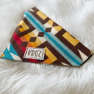 Pode incluir: Bandana triangular para animais de estimação com um padrão geométrico em tons de castanho, amarelo, azul-petróleo e vermelho. A bandana tem um patch bordado branco que diz "KRUZ".
