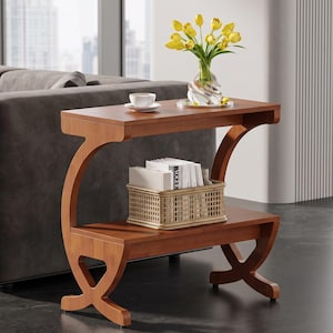 May include: A brown wooden side table with a rectangular top and a lower shelf. The table features curved, C-shaped supports on both sides. A white teacup, a glass vase with yellow tulips, and a woven basket containing books sit on the table.