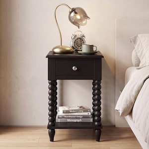 May include: A black wooden nightstand featuring a single drawer with a silver knob and four spindle-style legs with a stacked ball design. The piece includes an open lower shelf holding three stacked books and a flat rectangular top surface.