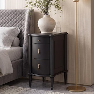 May include: A black bedside table with three drawers, featuring a small top drawer with a knob and two larger lower drawers with vertical drop handles. The piece stands on four tapered legs and is positioned next to a bed and a floor lamp.