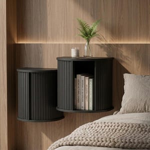 May include: Two matte black wall-mounted bedside cabinets with vertical slatted tambour doors. One cabinet is closed, while the other is open, revealing a shelf containing four books. A glass vase with a green plant and a candle sit on top.