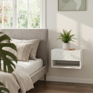 May include: A white floating nightstand featuring a single drawer with a diamond-patterned front and an open storage shelf underneath. The nightstand is mounted on a wall next to a bed with a gray headboard and a green potted plant on top.