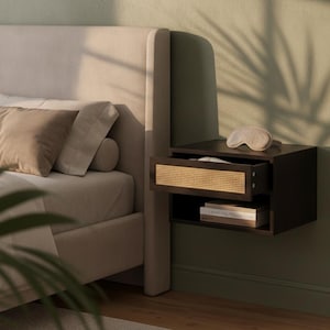May include: A dark brown floating nightstand featuring a single drawer with a light tan woven rattan front panel. The nightstand is mounted to a wall and includes an open storage shelf below the drawer, currently holding a book and a sleep mask.