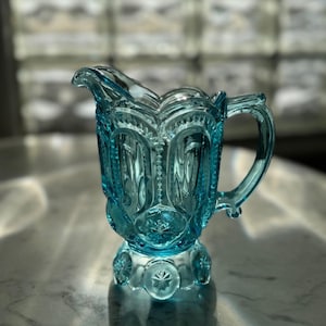 Puede incluir: Una jarra de crema de vidrio azul claro vintage con un patrón decorativo. La jarra de crema presenta un asa, un pico y un diseño de estrella en la base. El vidrio es transparente y está sobre una superficie de mármol.