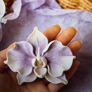 May include: A handcrafted orchid flower in shades of lavender and white, with a detailed center. The flower is held in a hand, showcasing its delicate petals and intricate design. The background is a soft purple fabric.
