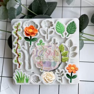 May include: A white silicone mold with various floral and leaf designs. The mold includes detailed shapes of flowers, leaves, and vines, with some designs colored in orange, green, and white. The text "Lil Stef's baking stuff Etsy" is visible.