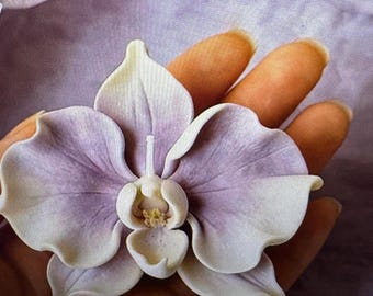 May include: A sculpted orchid flower ornament featuring soft lavender and white petals with a central column and labellum. The flower has a smooth, matte finish with delicate, wavy edges on each petal, mimicking the natural form of a phalaenopsis.