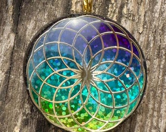 May include: A round pendant with a gold-colored bail. The pendant features a geometric design with a gradient of colors, including blue, purple, and green, and is filled with glitter. The pendant is set against a wooden background.