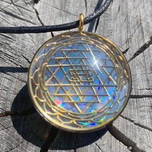 Puede incluir: Un collar con un colgante de oro redondo de vidrio transparente que contiene un símbolo Sri Yantra dorado. El colgante está en un cordón negro.