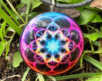 May include: A round pendant with a vibrant, colorful floral design in shades of blue, pink, and orange. The pendant hangs from a black cord necklace. The pendant is set against a backdrop of green foliage and brown earth.
