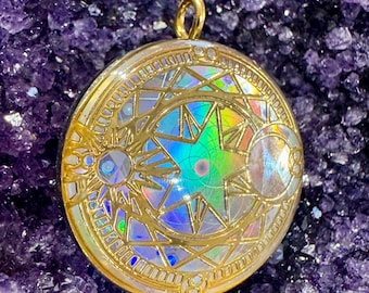 May include: A round, gold-colored pendant with an iridescent star and geometric design. The pendant has a gold loop at the top for hanging. The background is a deep purple, crystalline surface.