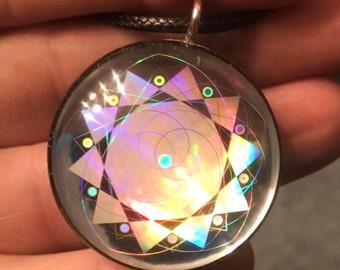 May include: A silver pendant with a clear glass dome containing a holographic geometric design. The design features a central spiral surrounded by a starburst pattern with black lines and iridescent colors.