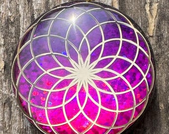 May include: A round pendant with a gold-colored bail. The pendant features a geometric design in silver over a gradient of purple and pink. The design has a flower-like shape in the center.