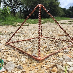 Könnte beinhalten: Eine kupferfarbene Pyramide aus verdrilltem Metalldraht, die auf einer steinigen Oberfläche ruht. Die Pyramide hat eine quadratische Basis und vier dreieckige Seiten mit einem detaillierten, strukturierten Aussehen. Der Hintergrund zeigt Grün und einen bewölkten Himmel.