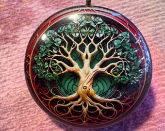 May include: Round pendant featuring a detailed tree of life design. The pendant has a silver-colored loop at the top. The tree has a tan trunk and branches with green leaves, set against a green and black background, framed by a red border.