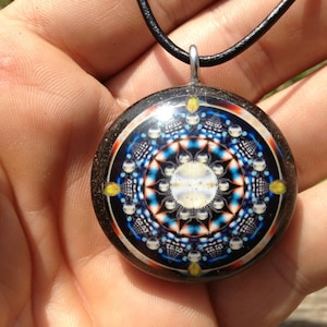 Könnte beinhalten: Eine runde Anhänger-Halskette mit einem schwarzen Band. Der Anhänger zeigt ein farbenfrohes Mandala-Design mit einem weißen Zentrum und einem schwarzen Hintergrund.