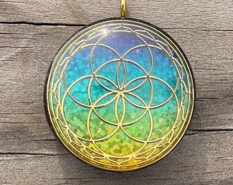 May include: A round pendant with a gold-colored geometric design overlaying a colorful background of blue, green, and yellow. The pendant has a gold-colored loop at the top for hanging. The background has a glittery effect.