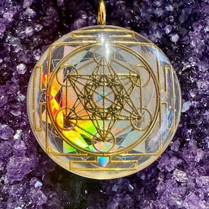 May include: A gold pendant with a clear resin dome featuring a geometric design. The design is a combination of a star of David, a flower of life, and a sacred geometry pattern. The pendant is set against a background of purple amethyst crystals.